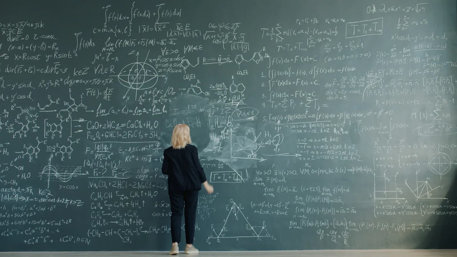 A person writing complex equations and physics formulas on a chalkboard wall